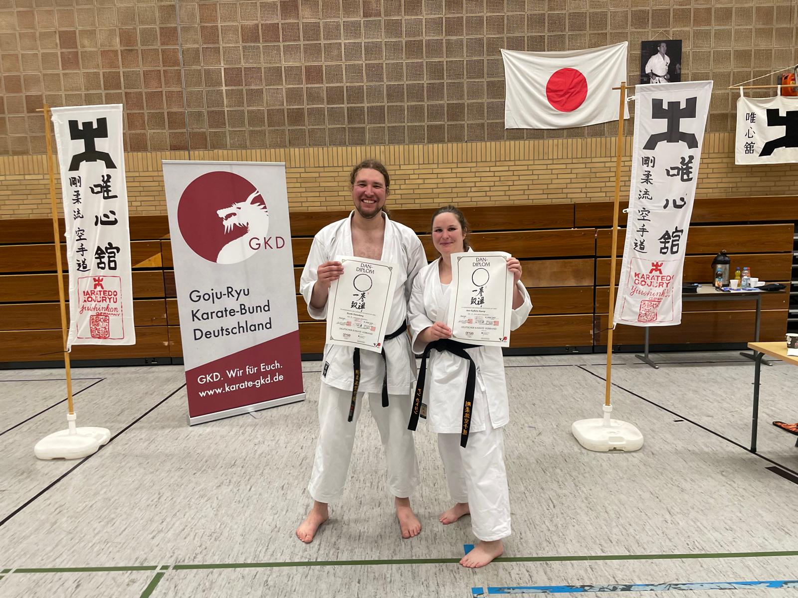 Ein junger Mann und eine Junge Frau halten eine Urkunden und freuen sich über eine bestandene Karate Prüfung.